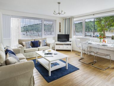 A living room with two beige sofas a white coffee table a TV on a stand and a dining table with transparent chairs by large windows at Leeside Dartmouth