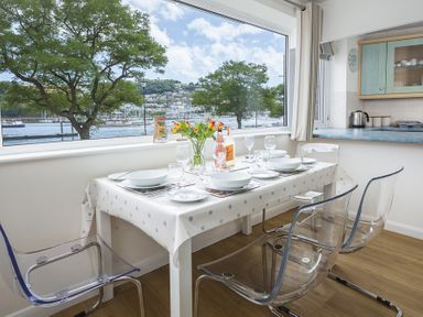 A dining area with a table set for four and clear chairs by a large window overlooking a waterfront at Leeside in Dartmouth
