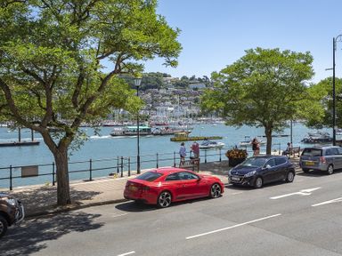 A waterfront street with parked cars and trees overlooking a harbor with boats and hills in the background at Leeside in Dartmouth