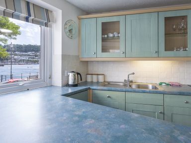 A kitchen with blue countertops green cupboards a stainless steel sink kettle and a window with a view of a marina at Leeside in Dartmouth