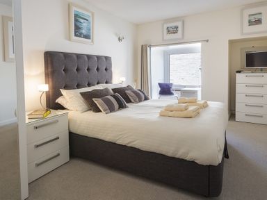 A bedroom with a bed with pillows and towels bedside tables with lamps a chair near the window and a chest of drawers with a television at Leeside in Dartmouth