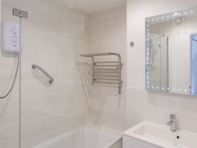 A bathroom with a bathtub shower combination a glass shower screen a wall-mounted electric shower a towel rack a mirror with lights and a sink at Leeside in Dartmouth