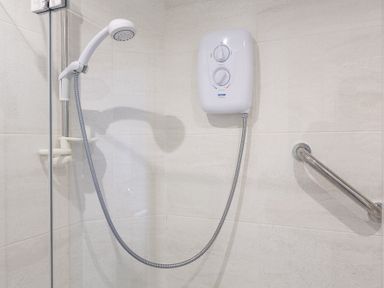 A shower area with a handheld showerhead electric water heater and a grab bar on tiled walls at Leeside Dartmouth