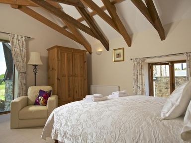A bedroom with wooden beams a wooden wardrobe an armchair and windows with curtains at Meadow View in Modbury