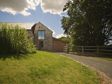 A stone house with a wooden extension along a gravel driveway and a lawn with plants and a wooden fence at Meadow View in Modbury