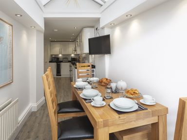 A dining area with a wooden table set with white dishes and croissants and a kitchen in the background at The Old Wash House in Dartmouth