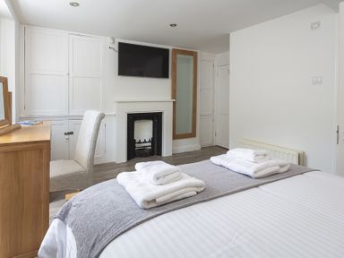 A bedroom with a bed with folded towels a fireplace a wall-mounted television a wooden desk with a chair and a tall mirror at The Old Wash House in Dartmouth