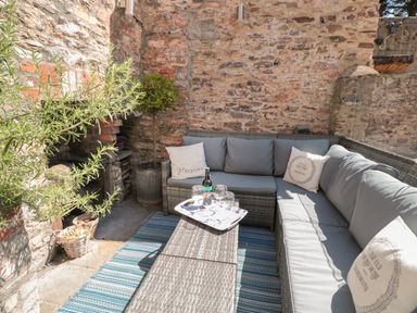 An outdoor seating area with a grey wicker sofa and cushions around a table with glasses and a bottle at The Old Wash House in Dartmouth