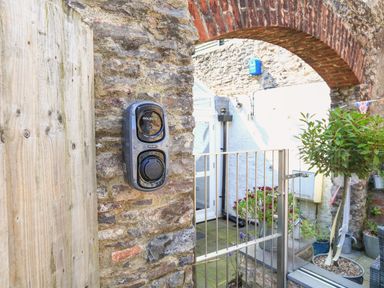 An outdoor area with a stone wall and wooden gate an electric vehicle charger mounted on the wall a metal gate a brick archway and various potted plants at The Old Wash House in Dartmouth