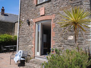 A stone cottage exterior with a door open to a dining area and outdoor chairs and plants at Orchard Barn in Salcombe