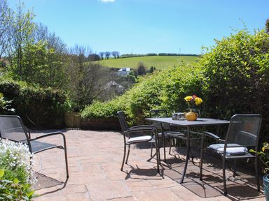 An outdoor patio with a metal table and four chairs with cushions overlooking a green hillside at Orchard Barn in Salcombe