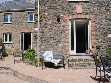 An outdoor patio area with metal chairs and a bench with a blanket against a stone building at Orchard Barn in Salcombe