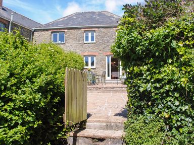 A stone house with a slate roof and four windows viewed through an open wooden gate with bushes on either side at Orchard Barn in Salcombe