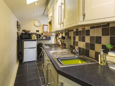 A narrow kitchen with tiled floor and backsplash a sink stove microwave small fridge and wall clock at Pixie Cottage in Dartmouth