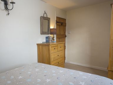 A bedroom with a wooden chest of drawers, a wicker framed mirror, a lamp, and a door at Pixie Cottage in Dartmouth