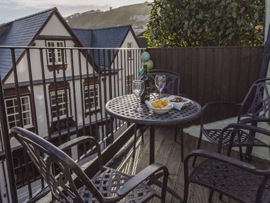 A balcony with a round metal table and four metal chairs with snacks and drinks with a view of houses and a hillside at Pixie Cottage in Dartmouth