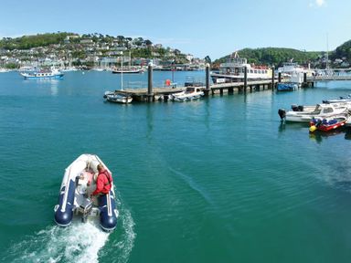 A small motorboat on water approaching a dock with several boats and a town on a hillside in the background at Pixie Cottage in Dartmouth