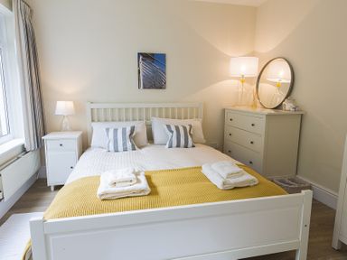 A bedroom with a white bed with striped pillows a yellow throw a bedside table and a dresser with a lamp and round mirror at Prospects in Salcombe