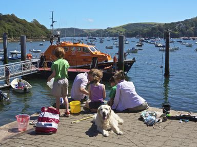 A group of people with a dog sitting and standing on a dock near boats and water at Prospects in Salcombe