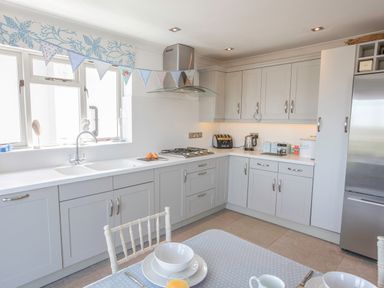 A kitchen with white cabinets a gas stove a stainless steel refrigerator and a dining table with plates and bowls at Raleigh House in Salcombe