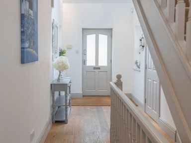 A hallway with a gray door wooden floor a small table with flowers a white railing and a painting on the wall at Raleigh House in Salcombe