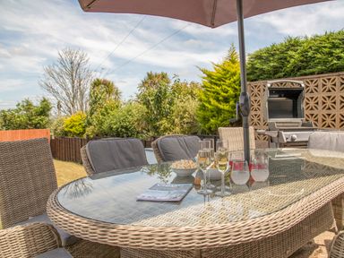 An outdoor patio with a glass table and wicker chairs under an umbrella with drinks and snacks with a barbecue grill in the background at Raleigh House in Salcombe