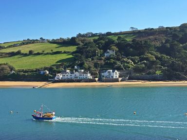 A boat moving on water near a sandy beach with houses and green hills in the background at Rock Point in Salcombe