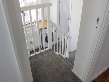 A stairway with grey carpet leading down to a utility area with a washing machine a sink and wooden countertop at Rock Point in Salcombe