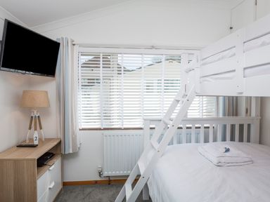 A bedroom with a white bunk bed folded towels on the bottom bed a wooden cabinet with a lamp and a wall-mounted television at Rock Point in Salcombe