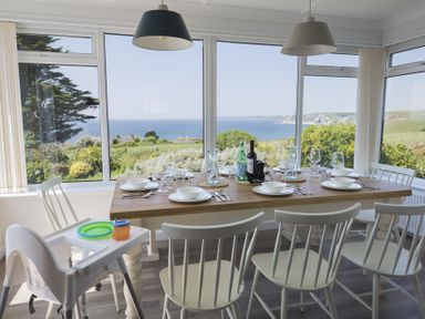 A dining room with a wooden table set for six people next to large windows showing a sea view at Seaspray in Bigbury-on-Sea