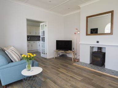 A living room with a blue armchair, small white table with yellow flowers, television on a wooden stand, wood stove in a tiled fireplace, and an open door to a kitchen at Seaspray Bigbury-on-Sea