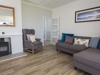 A living room with a gray armchair and sectional sofa with cushions next to a fireplace and a wall mirror at Seaspray Bigbury-on-Sea