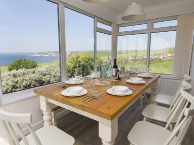 A dining area with a wooden table set with plates and glasses surrounded by white chairs with large windows showing a coastal view at Seaspray Bigbury-on-Sea