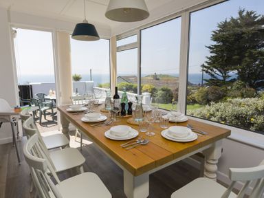 A dining room with a wooden table set with plates glasses and cutlery and white chairs by large windows overlooking a garden and sea at Seaspray Bigbury-on-Sea