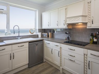 A kitchen with white cabinets a sink a dishwasher and a window showing a sea view at Seaspray Bigbury-on-Sea