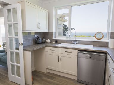 A kitchen with white cabinets a sink dishwasher kettle and a window with sea view at Seaspray Bigbury-on-Sea