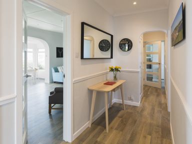A hallway with a wooden table holding a vase of yellow flowers and a book a mirror and clock on the wall and doors leading to other rooms at Seaspray Bigbury-on-Sea