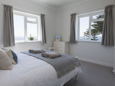 A bedroom with a bed, pillows, folded towels, a chest of drawers, stool, and windows with curtains at Seaspray Bigbury-on-Sea