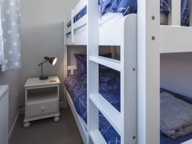 A bedroom with a white bunk bed blue bedding a white nightstand with a lamp and a window with star patterned curtains at Seaspray Bigbury-on-Sea