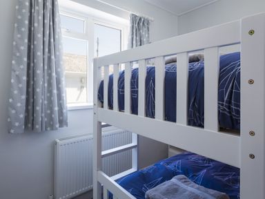 A bedroom with white bunk beds and blue bedding with sailboat patterns near a window with star-patterned curtains at Seaspray Bigbury-on-Sea in Bigbury-on-Sea