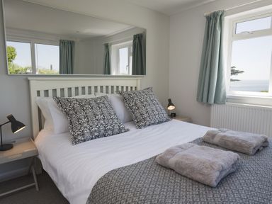 A bedroom with a white bed frame, patterned pillows, folded towels on a blanket, bedside tables, lamps, and windows with curtains at Seaspray Bigbury-on-Sea