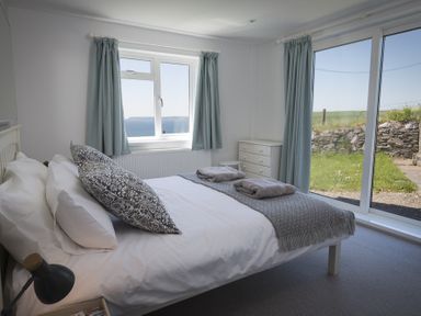 A bedroom with a bed with white and patterned pillows folded towels on a blanket and two windows with curtains at Seaspray Bigbury on Sea