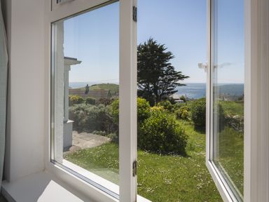 A view through an open window showing a garden with bushes and a tree and the sea in the distance at Seaspray Bigbury-on-Sea