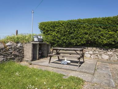 An outdoor patio area with a picnic table and a built-in brick barbecue against a hedge at Seaspray Bigbury-on-Sea