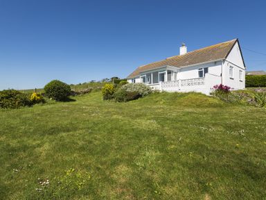 A white house with a sloped roof on a grassy hill with bushes and flowers at Seaspray Bigbury-on-Sea