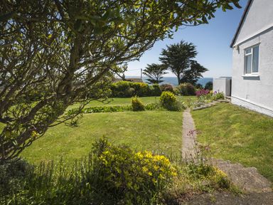 A garden with grass, bushes, a tree, and a pathway beside a white house with the sea in the background at Seaspray Bigbury-on-Sea