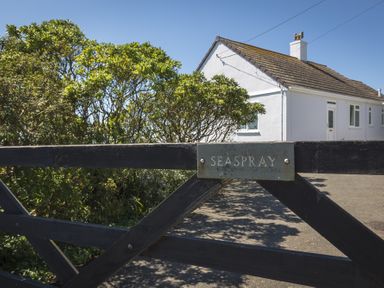 A black wooden gate with a nameplate reading seaspray in front of a white house and green bushes at Seaspray Bigbury-on-Sea