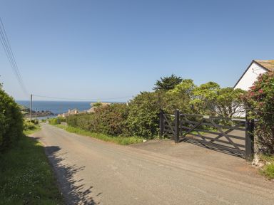 A narrow road with hedges and a black wooden gate leading to a driveway with a view of the sea at Seaspray Bigbury-on-Sea