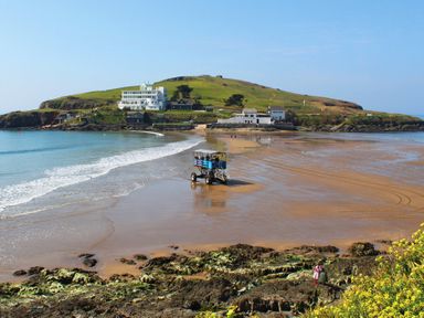 A sandy beach with a vehicle carrying people and an island with buildings and greenery in the background at Seaspray Bigbury on Sea