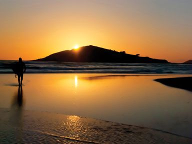 A person carrying a surfboard walking on a beach during sunset at Seaspray Bigbury-on-Sea